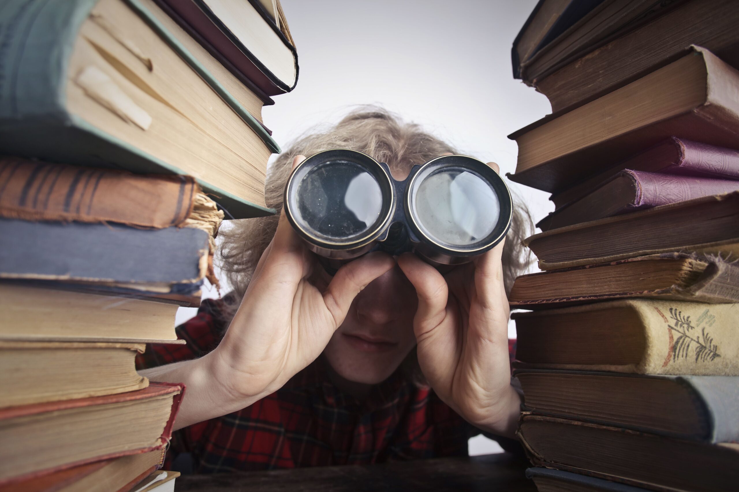 Young unrecognizable person sitting at table with stacked old books and looking through binoculars