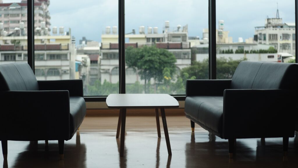 Contemporary lounge with two sofas and a table, overlooking an urban cityscape through large windows.