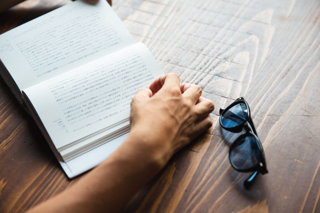 High angle crop anonymous person sitting at table with stylish dark blue sunglasses and reading interesting book
