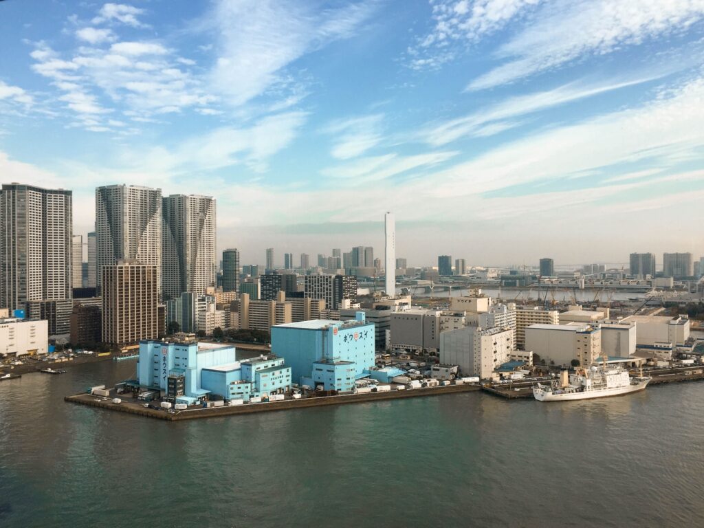 【2026年最新】工場の無人化事例5選!導入コスト・削減効果と失敗しない進め方 Aerial view of a modern city skyline with skyscrapers and waterfront buildings under a clear blue sky.