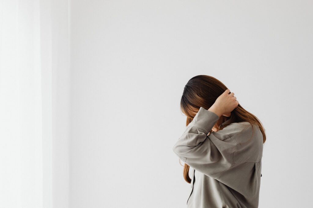 A minimalist portrait of a woman indoors with her hands in her hair, featuring a white wall.