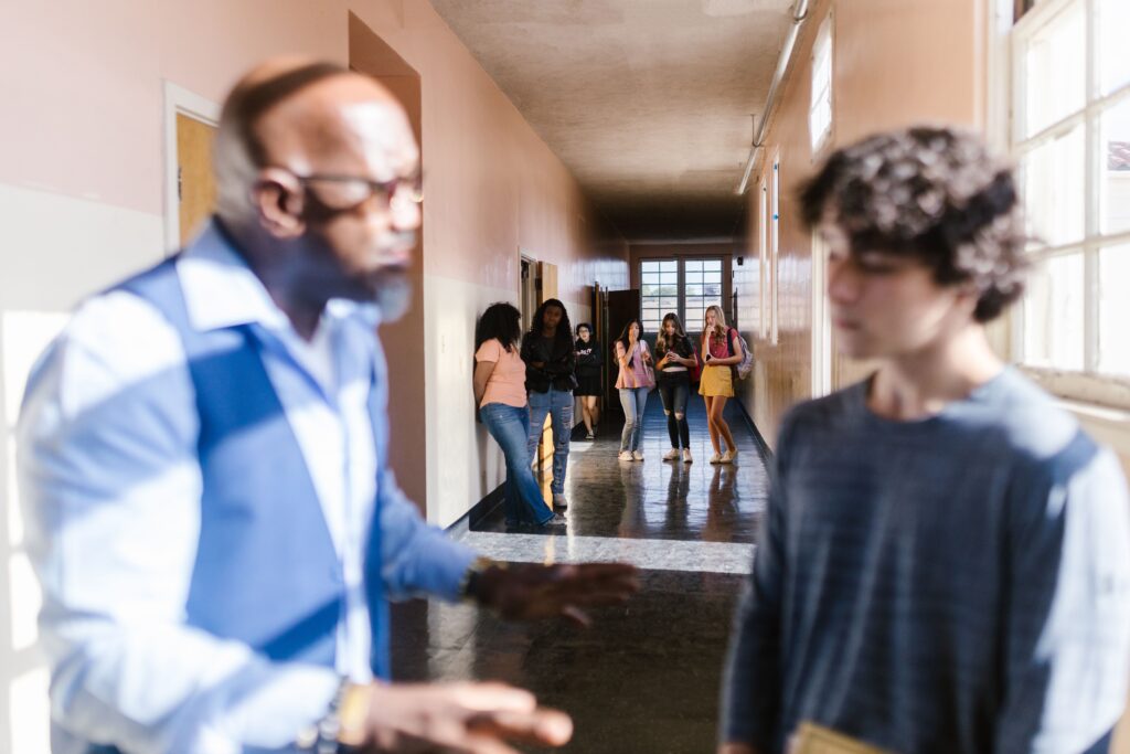 A blurred teacher talks to a student in a school hallway, with peers in the background.